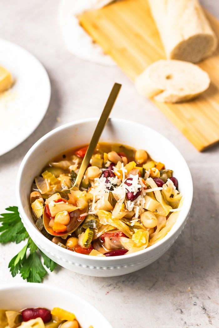 A white bowl with minestrone soup with shredded parmesan on top and a gold spoon. 
