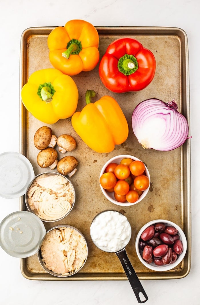Bell peppers, a red onion half, mushrooms, cans of tuna, a measuring cup with cottage cheese, cherry tomatoes and kalamata olives on a baking sheet.
