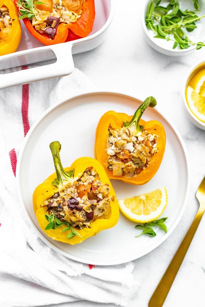 A yellow and orange stuffed bell pepper with a Greek tuna stuffing on a plate and garnished with herbs and a lemon wedge.