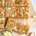 Apple granola bars on a wood board next to a bowl of white chocolate chips and apple slices.