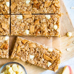 Apple granola bars on a wood board next to a bowl of white chocolate chips and apple slices.