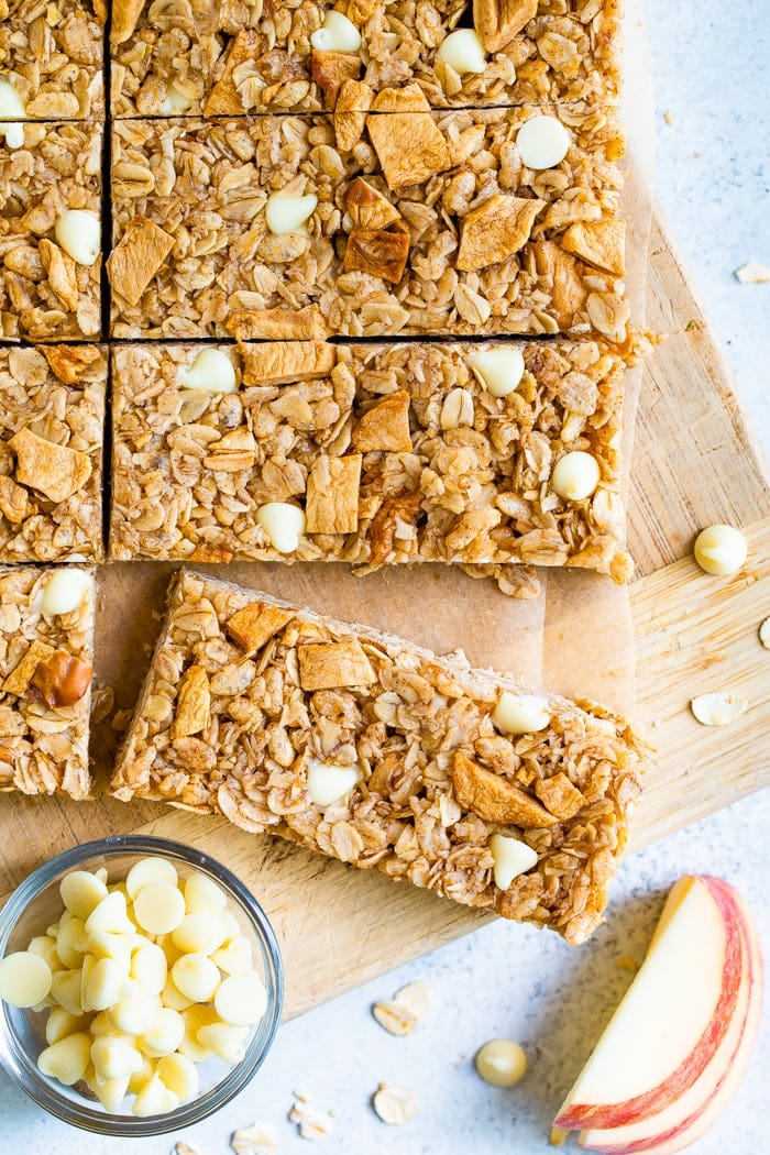 Apple granola bars on a wood board next to a bowl of white chocolate chips and apple slices.