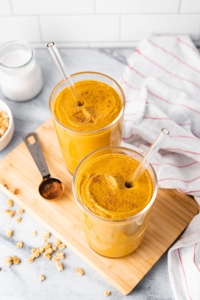 Two glasses with thick pumpkin smoothie and two straws. A teaspoon with pumpkin spice is next to the glasses.