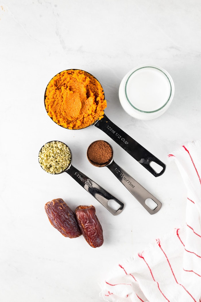 Dates, hemp seeds, pumpkin pie spice, pumpkin and coconut milk measured out on a table next to a striped kitchen towel.