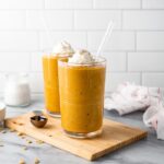 Two pumpkin pie smoothies on a wood cutting board surrounded by spices and a kitchen towlel.