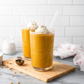 Two pumpkin pie smoothies on a wood cutting board surrounded by spices and a kitchen towlel.