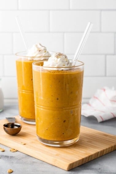 Two pumpkin pie smoothies on a wood cutting board surrounded by spices and a kitchen towlel.