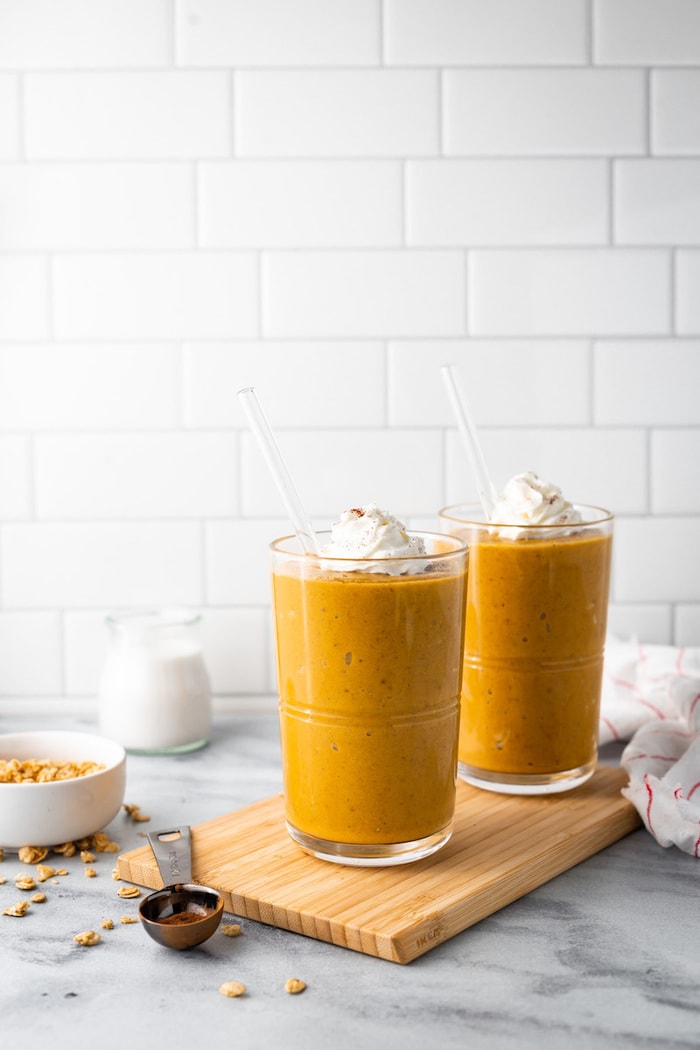 Two pumpkin pie smoothies on a wood cutting board surrounded by spices and a kitchen towlel.