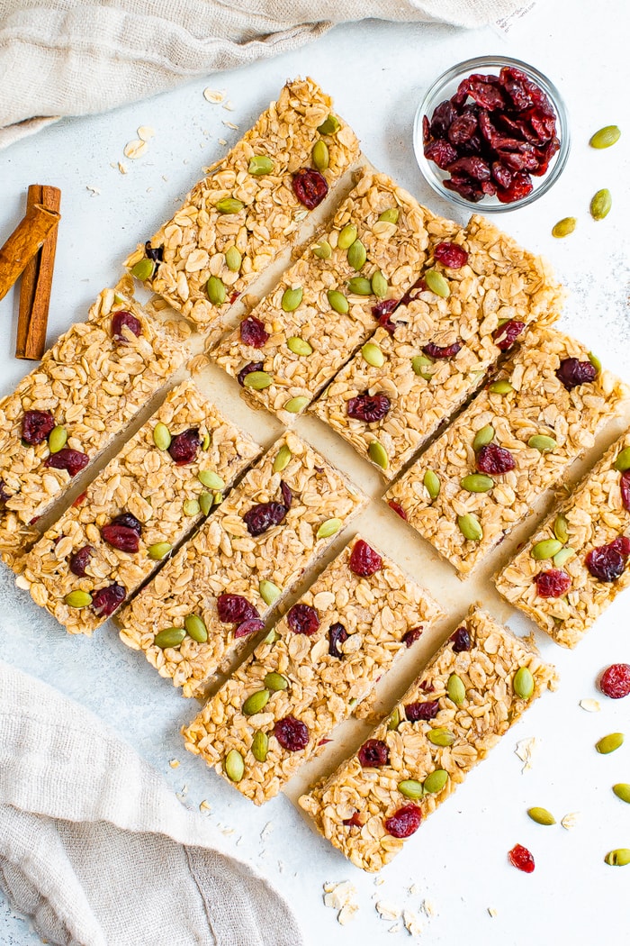 Pumpkin spice granola bars neatly on a table surrounded by pepitas, cranberries and cinnamon.
