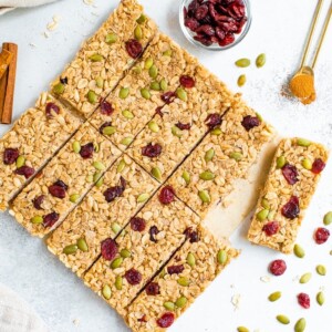 Pumpkin spice granola bars neatly on a table surrounded by pepitas, cranberries and cinnamon.