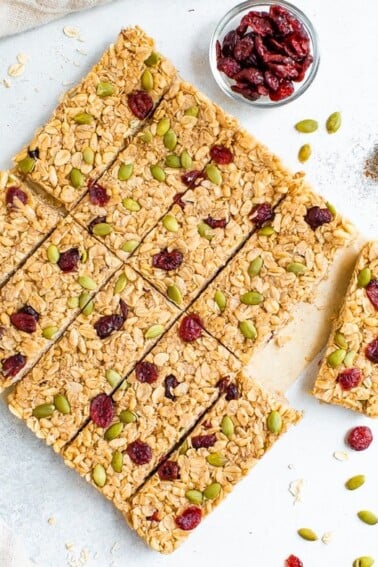 Pumpkin spice granola bars neatly on a table surrounded by pepitas, cranberries and cinnamon.