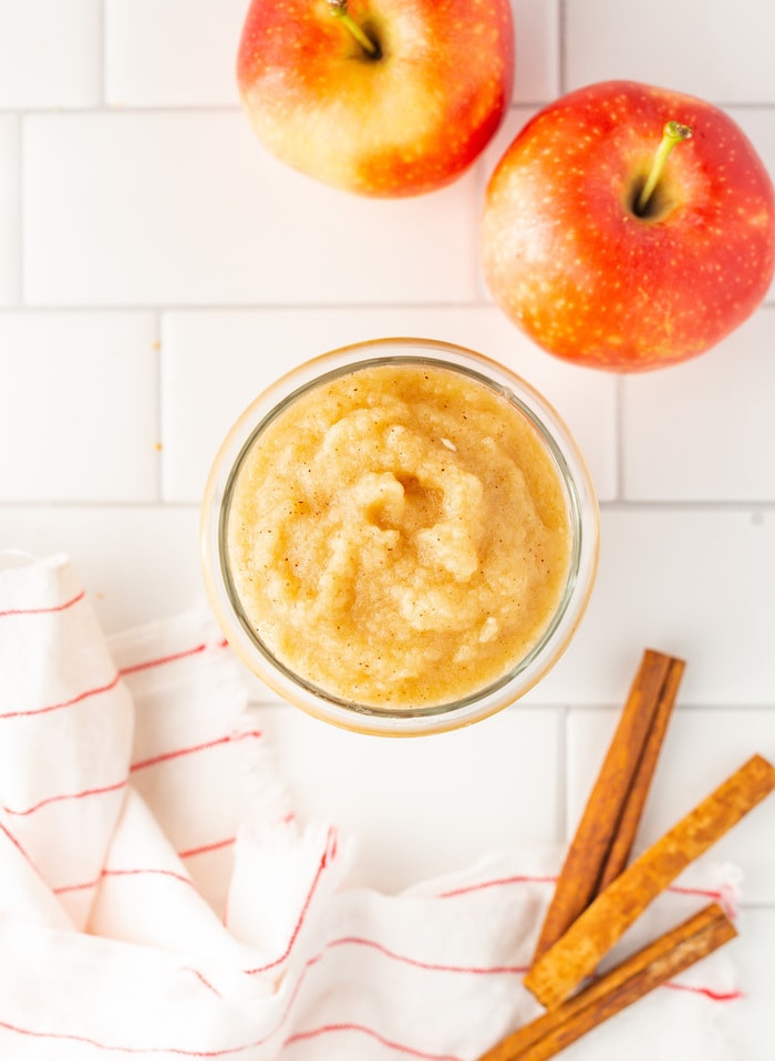 Bird's eye view of glass jar full of homemade applesauce. Surrounded by a cloth napkin, apples and cinnamon sticks.