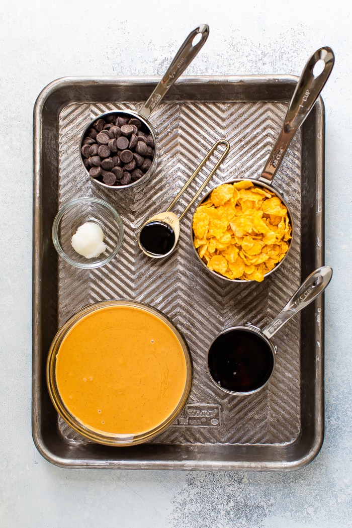 Overhead shot of baking sheet with ingredients measured out. Peanut butter, maple syrup, coconut oil, corn flakes and chocolate chips in measuring cups.