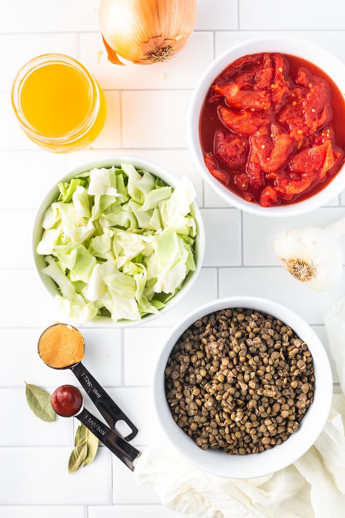 Ingredients needed for cabbage lentil soup with a white tile background.