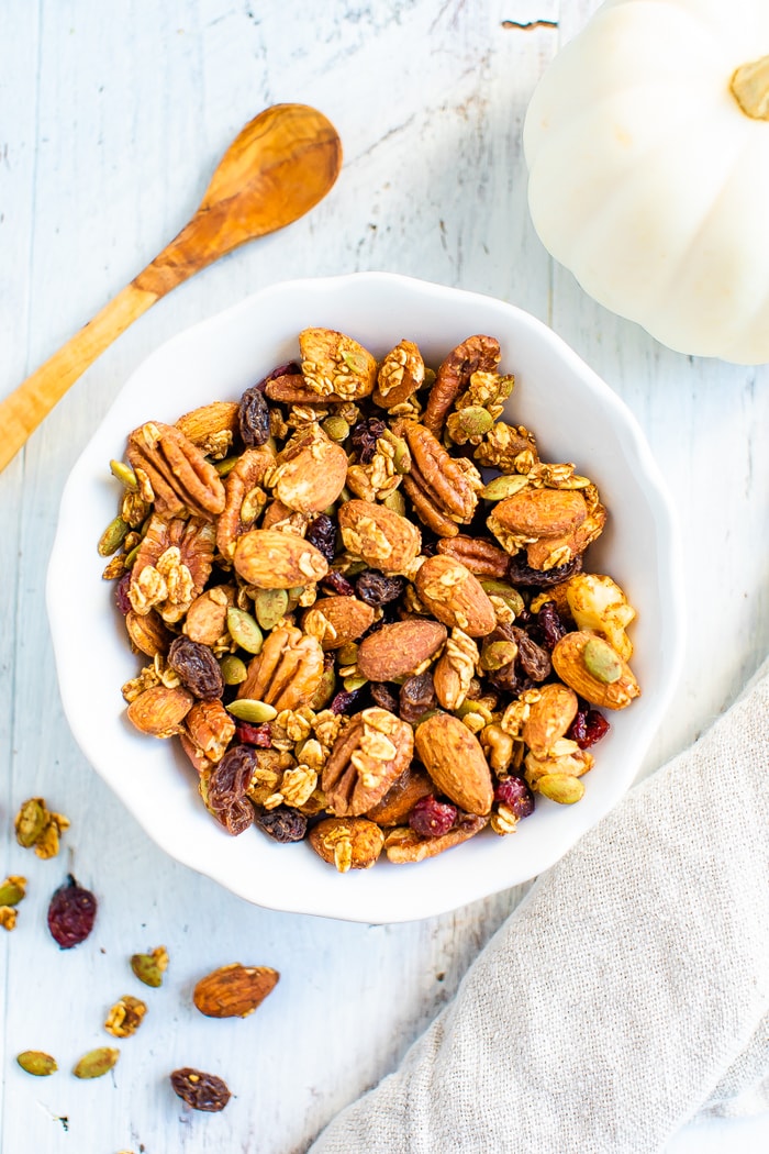 White bowl full of pumpkin spice trail mix made with granola, fruit and nuts. A small wooden spoon is beside the bowl.