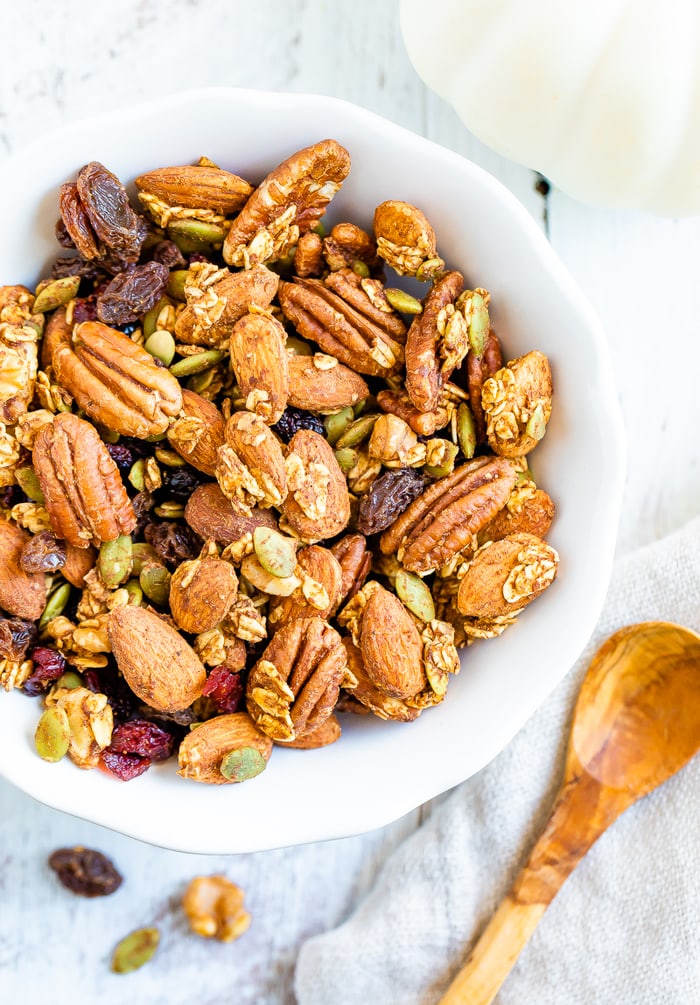 White bowl full of pumpkin spice trail mix made with granola, fruit and nuts. A small wooden spoon is beside the bowl.