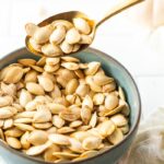 Spoon scooping up some roasted pumpkin seeds from a bowl.