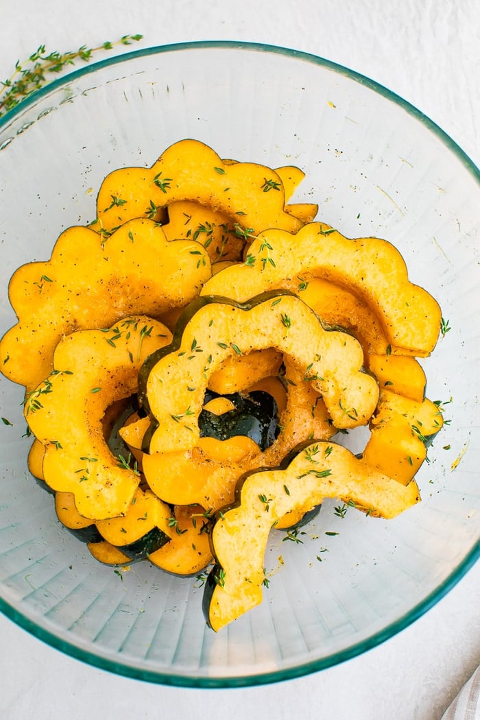 Acorn squash slices in a bowl with herbs, salt and pepper.