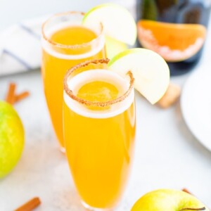 Two tall glasses of apple cider mimosas with a cinnamon sugar rim and an apple slice for garnish.