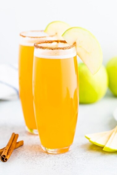 Two tall glasses of apple cider mimosas with a cinnamon sugar rim and an apple slice for garnish.