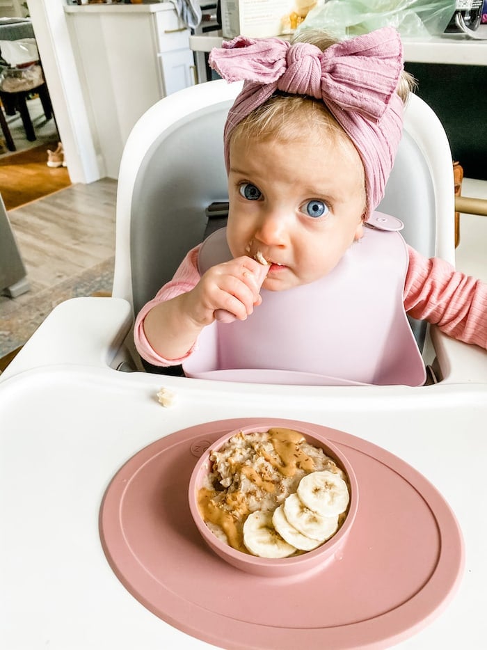 Baby Olivia in a highchair eating a bowl of oatmeal with bananas and peanut butter on top.