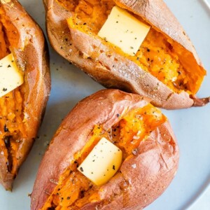 Three baked sweet potatoes on a plate topped with butter, salt and pepper.