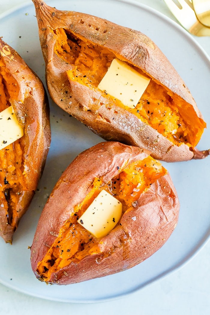 Three baked sweet potatoes on a plate topped with butter, salt and pepper.