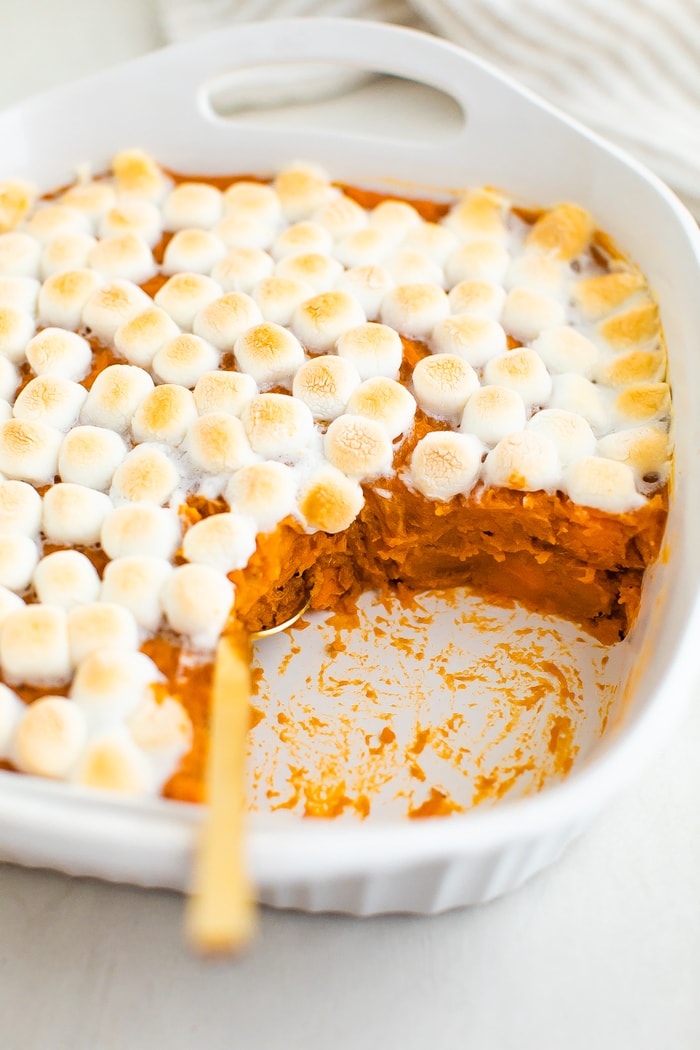 Sweet potato casserole topped with marshmallows. A spoon is in the casserole scooping a portion out.