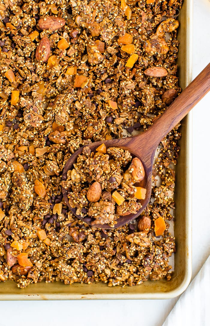 Wood spoon scooping up quinoa granola made with chocolate chips, nuts and dried fruit. Granola is on a baking sheet.