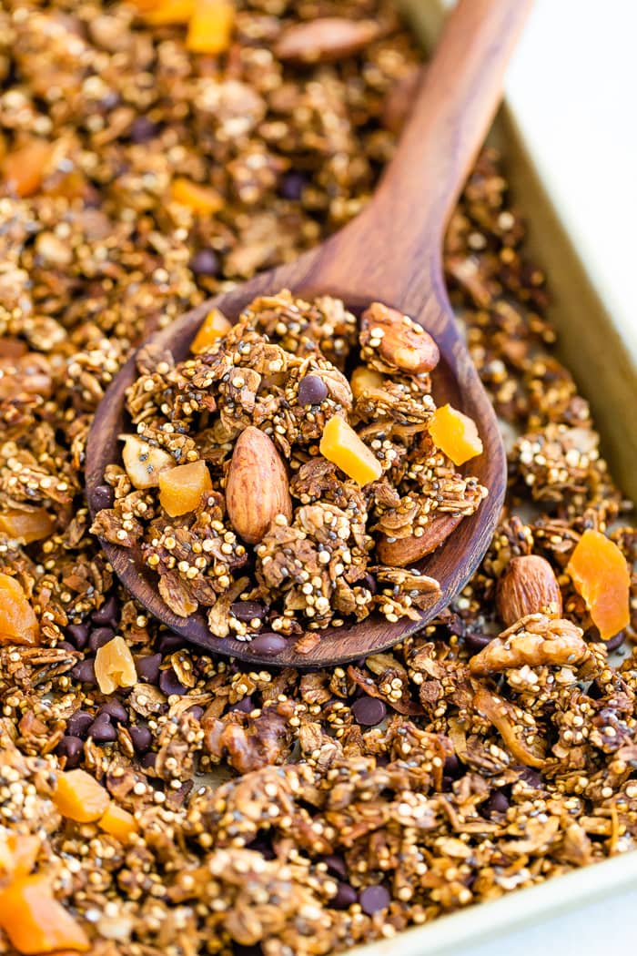 Wood spoon scooping up quinoa granola made with chocolate chips, nuts and dried fruit. Granola is on a baking sheet.