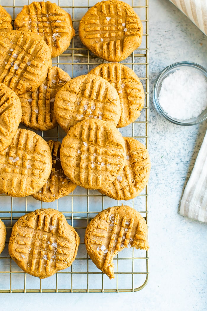 Peanut butter cookies topped with flakey sea salt on a cooling rack. One cookie has a bite out of it. A small bowl of sea salt is beside the rack.