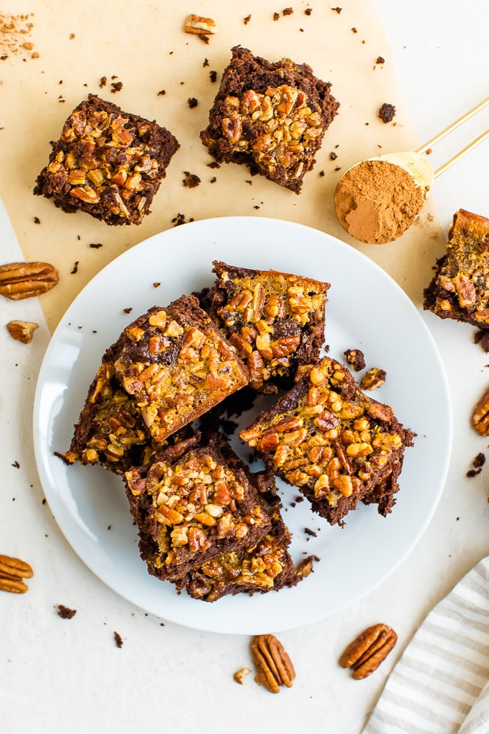 Pecan pie brownies on a plate. Plate is surrounded by more brownies, a spoon with cocoa powder and pecans.