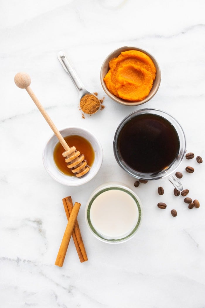 Ingredients to make a homemade pumpkin spice latte on a marble countertop: pumpkin, pumpkin spice, coffee, almond milk and maple syrup..