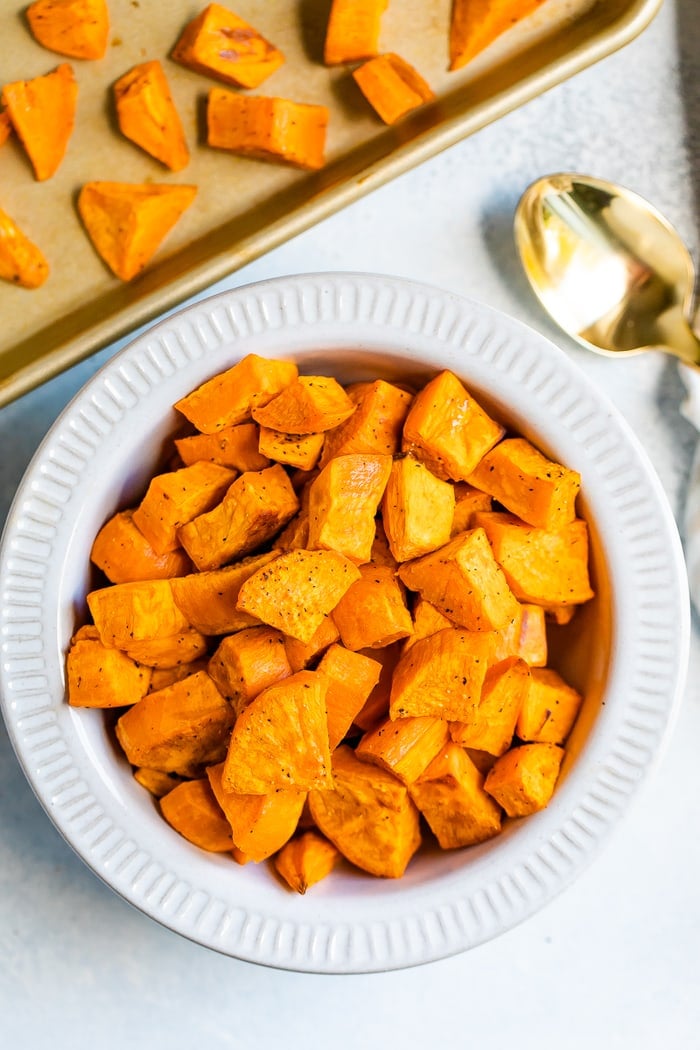 Bowl of roasted sweet potato chunks. A spoon and sheet pan with potatoes is beside the bowl.