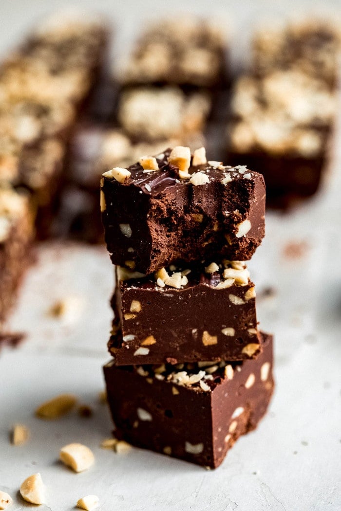 Stack of three squares of fudge studded with chopped peanuts. The top piece of fudge has a bite taken out of it.