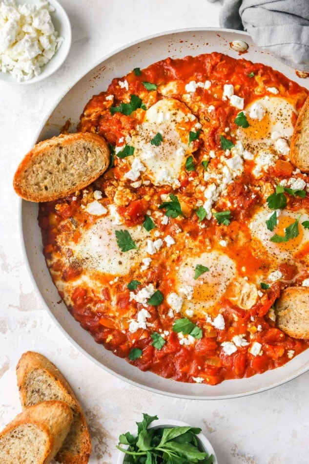 Cooked shakshuka in a sauté pan with fresh herbs, feta and a slice of crusty bread.