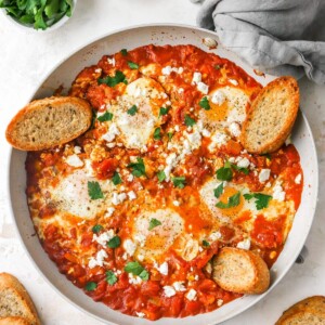 Cooked shakshuka in a sauté pan with fresh herbs, feta and slices of crusty bread.