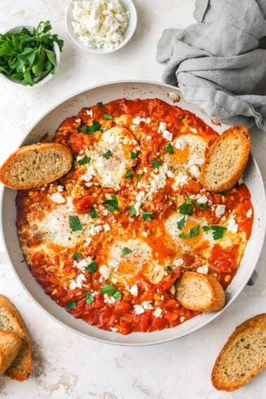 Cooked shakshuka in a sauté pan with fresh herbs, feta and slices of crusty bread.