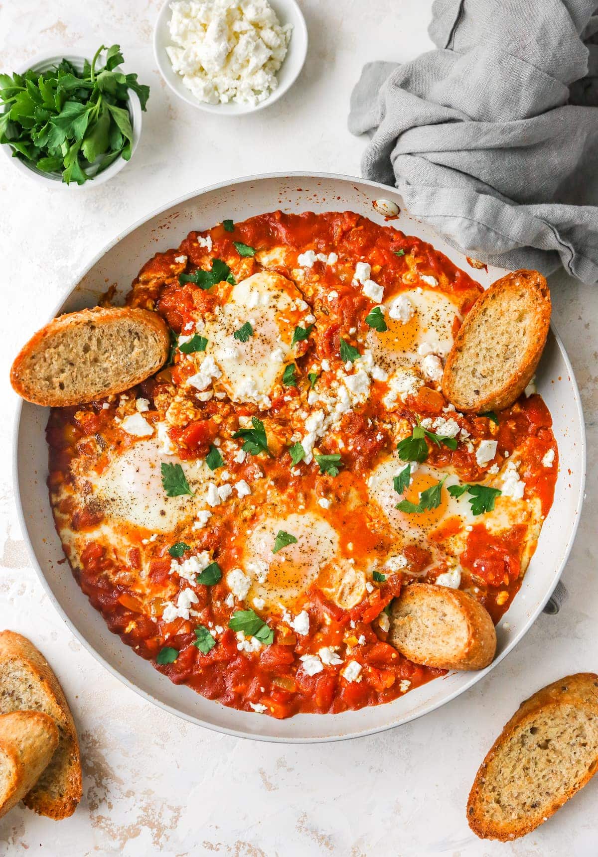 Cooked shakshuka in a sauté pan with fresh herbs, feta and slices of crusty bread.