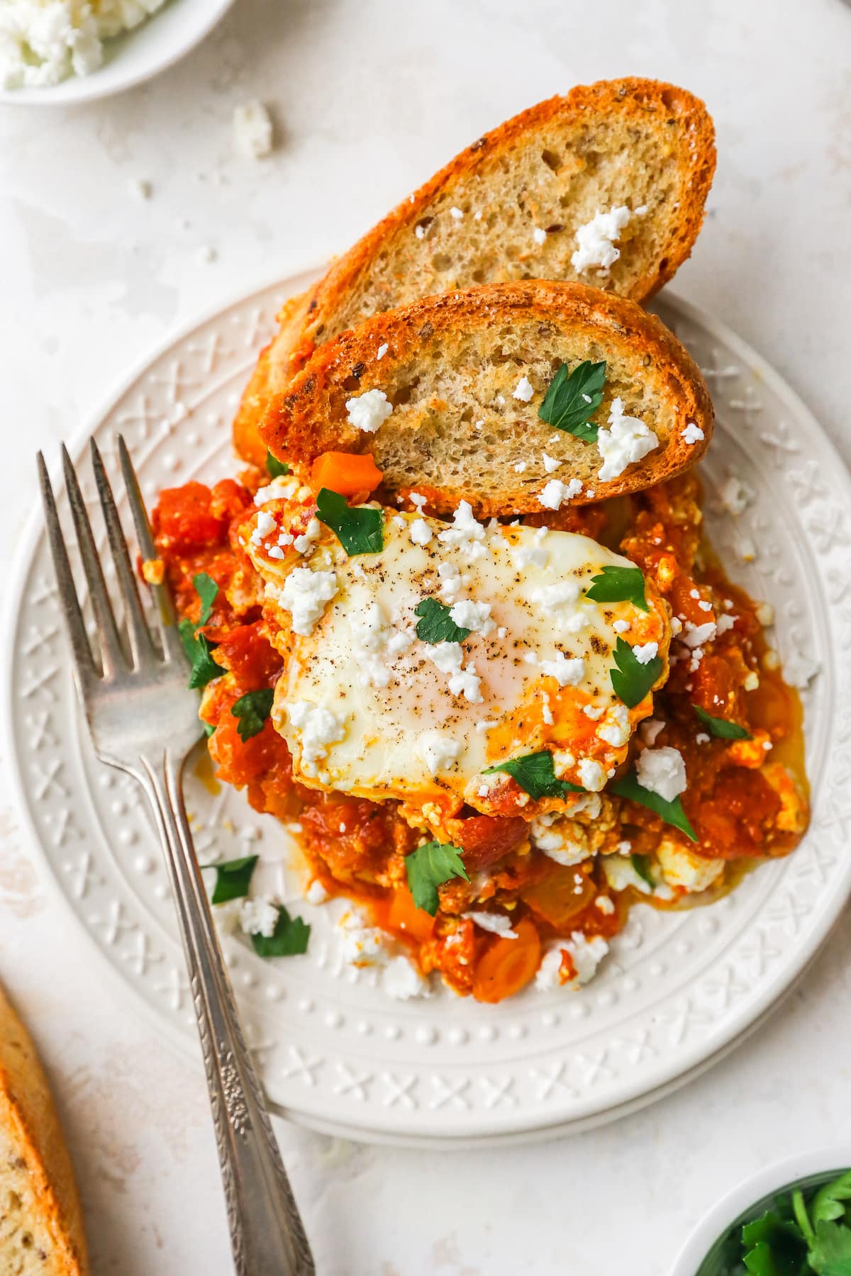 Plated shakshuka on a white plate with fresh herbs, feta and two slices of crusty bread.