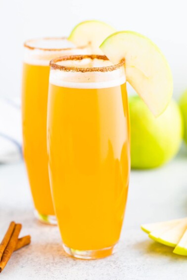 Two stemless champagne glasses of apple cider mimosas with a cinnamon sugar rim, cinnamon sticks and an apple slice for garnish.