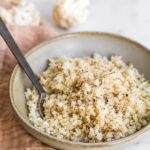 Bowl full of cauliflower rice. A spoon is in the bowl. A pink napkin is below the bowl.