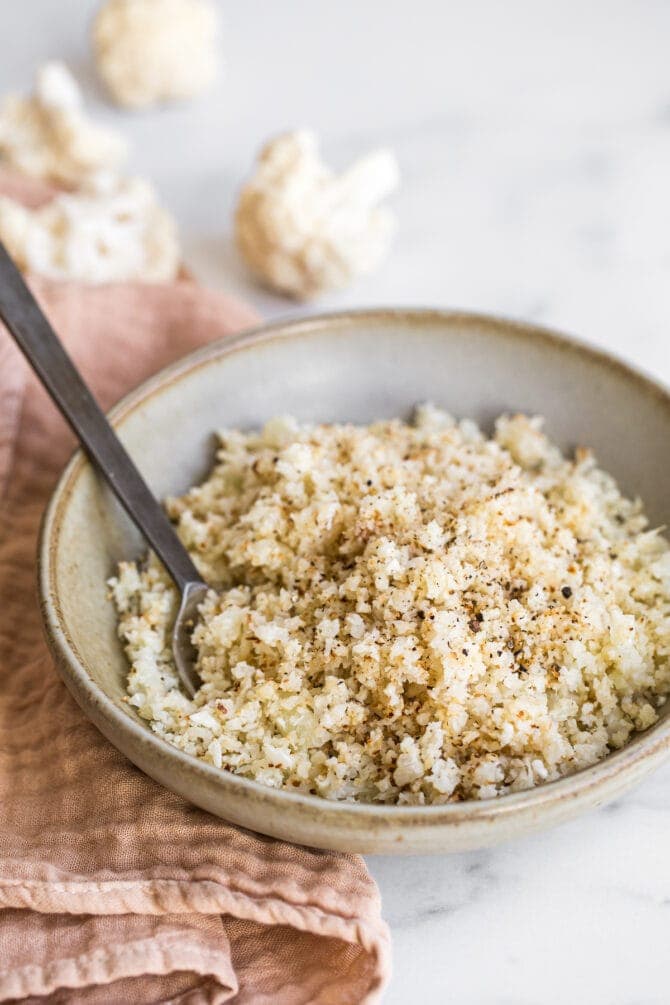 Bowl full of cauliflower rice. A spoon is in the bowl. A pink napkin is below the bowl.