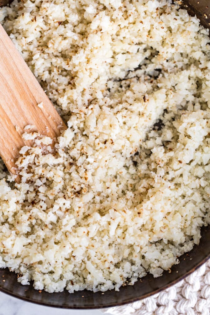 Cauliflower rice in a pan with a wooden spatula stirring it.