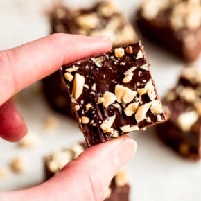 Square image with hand holding a piece of fudge topped with chopped peanuts.