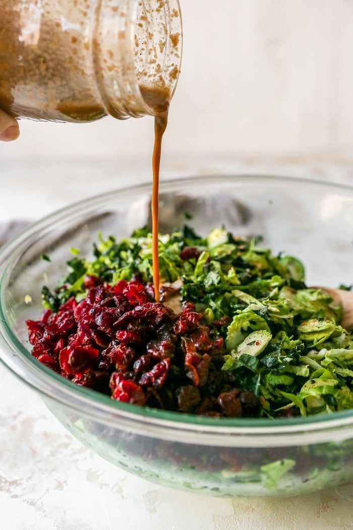 Homemade dressing being poured over a mixing bowl with shredded kale, brussles and dried cranberries.