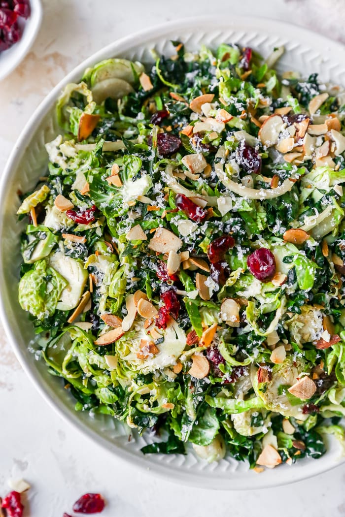 Kale and brussels sprout salad in a serving bowl topped with dried cranberries, almonds and cheese.