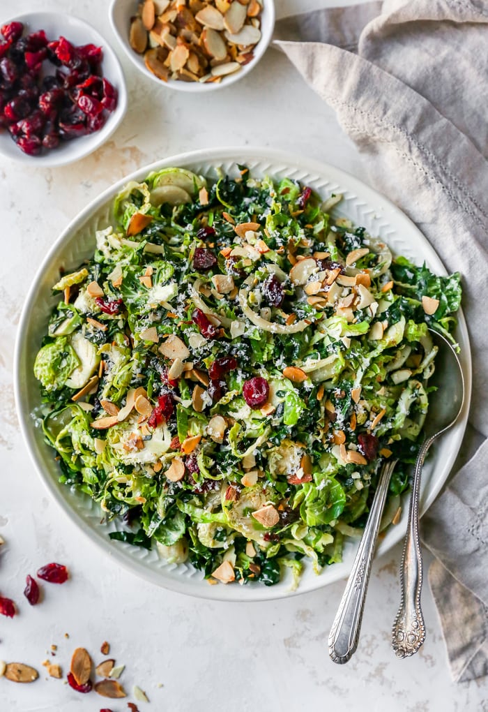Kale and brussels sprout salad in a bowl with serving spoons. A bowl of cranberries and almond are beside the salad bowl.