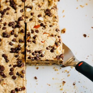 Spatula under a cookie bar in a baking pan. The cookie bar is topped with almonds and mini chocolate chips.