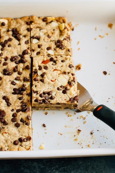 Spatula under a cookie bar in a baking pan. The cookie bar is topped with almonds and mini chocolate chips.